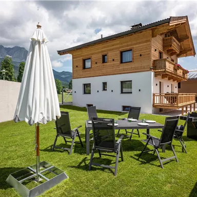 A modern wooden house with a beautiful garden and a view of the mountains. There is a table with chairs, a parasol, and a grill outside.