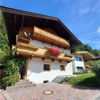 A charming house in alpine style with wooden balustrades and flower balconies. The sky is clear and blue, surrounded by lush greenery.