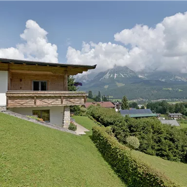 A beautiful house on a hill overlooking the mountains. The landscape is green and wooded, under a clear sky with some clouds.