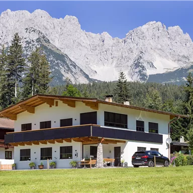 A modern house with a beautiful garden, surrounded by mountains and trees. The clear sky gives the scene a tranquil atmosphere.