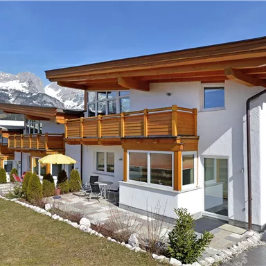 A modern house with wooden cladding and large windows. In the background, mountains and a blue sky can be seen.