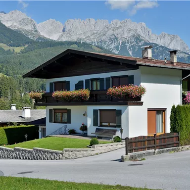 Ein schönes Haus in den Bergen mit blühenden Pflanzen auf dem Balkon. Im Hintergrund erstrecken sich majestätische Berge und ein klarer Himmel.