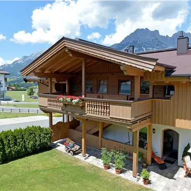 A cozy wooden house with a balcony and colorful flowers. In the background, the mountains and a clear sky are visible.