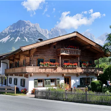 A traditional wooden house with a balcony full of flowers. In the background, majestic mountains and a blue sky can be seen.