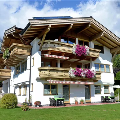Ein schönes Chalet im alpinen Stil mit Balkonen und bunten Blumen. Im Hintergrund sind Berge und ein klarer Himmel zu sehen.