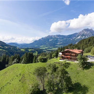 Eine malerische Landschaft mit sanften Hügeln und einem traditionellen Holzhaus. Im Hintergrund sind majestätische Berge und ein klarer, blauer Himmel zu sehen.