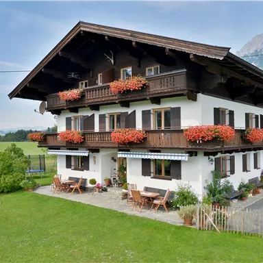 A traditional alpine house with flower boxes and a terrace. In the background, gentle hills and mountains can be seen.