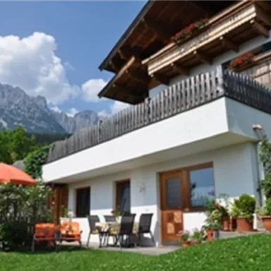 A charming house with a balcony and a terrace, surrounded by green grass and potted plants. In the background, majestic mountains and a clear blue sky can be seen.