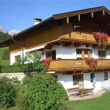 A cozy, traditional wooden house with blooming balconies. Surrounded by green meadows and mountains in the background.