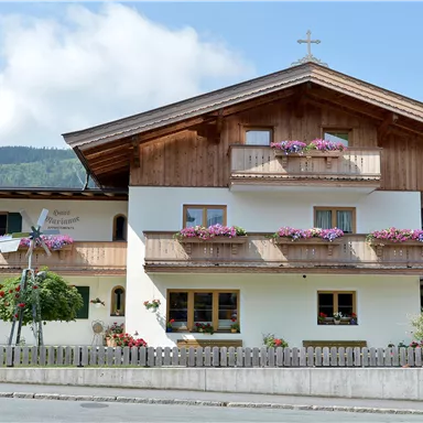 Ein traditionelles alpenländisches Haus mit Holzverkleidung und Blumenkästen. Im Hintergrund sind sanfte Hügel und ein blauer Himmel zu sehen.