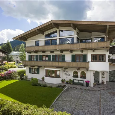 A modern house with a large balcony and a beautiful garden landscape. The surroundings are green and surrounded by mountains.
