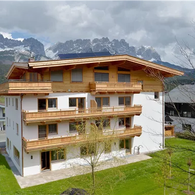 A modern house with wooden cladding and balconies, surrounded by green grass. In the background, impressive mountains and a cloudy sky can be seen.