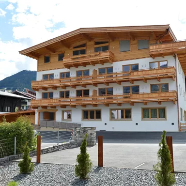 A modern building in alpine style with several balconies. Surrounded by a green landscape and mountains in the background.