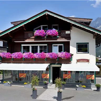 A beautiful old house with balconies, decorated with colorful flowers. In the background, mountains and a blue sky can be seen.