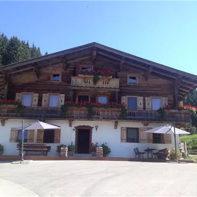 A traditional wooden house with balconies full of flowers. It is located in a picturesque rural setting with green hills.