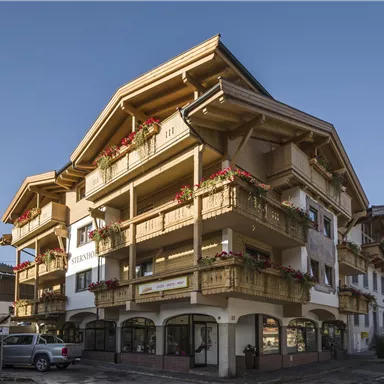 Ein charmantes Gebäude im alpinen Stil mit mehreren Balkonen und Blumen. Die klare Sicht auf die Berge im Hintergrund ergänzt die malerische Kulisse.