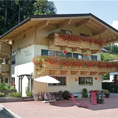 A cozy wooden house with blooming balconies and a sunny terrace. The surroundings are green and inviting.