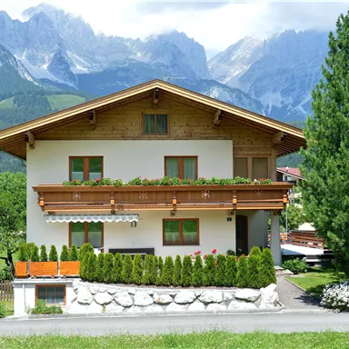 Ein charmantes Haus mit Holzveranda und blühenden Pflanzen. Im Hintergrund erstrecken sich majestätische Berge und ein klarer Himmel.