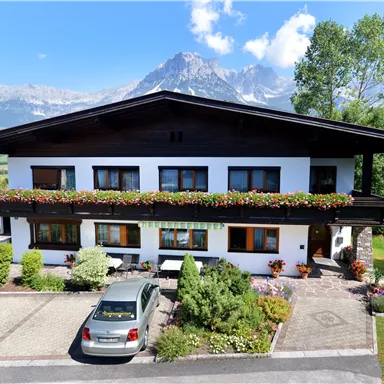 A charming house with flower windows and a carport. In the background, majestic mountains and a blue sky can be seen.