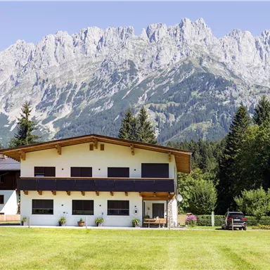 A modern house with a lawn and trees in the foreground. In the background, impressive mountains rise under a clear sky.