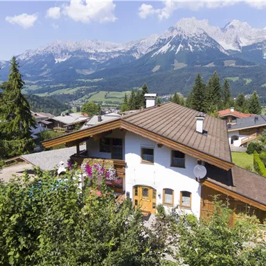 Ein gemütliches Haus inmitten von grünen Bäumen und Blumen. Im Hintergrund ragen majestätische Berge unter einem klaren Himmel empor.