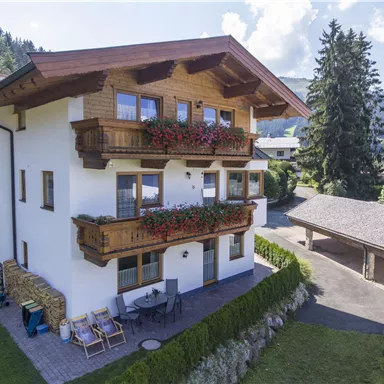Ein schönes, modernes Haus mit Holzverzierungen und Balkonen, umgeben von Natur. Im Vordergrund gibt es einen gepflegten Garten und eine überdachte Terrasse.