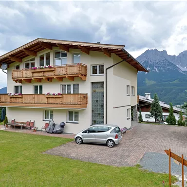 Ein modernes Haus mit Balkonen und einer grünen Rasenfläche. Im Hintergrund sind Berge unter einem bewölkten Himmel sichtbar.