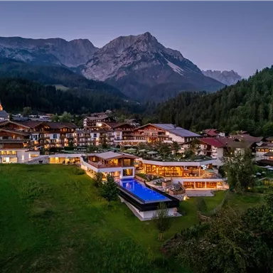 Ein malerisches Hotel in den Bergen mit modernem Design. Im Vordergrund sind ein Pool und grüne Wiesen zu sehen, umgeben von majestätischen Bergen.