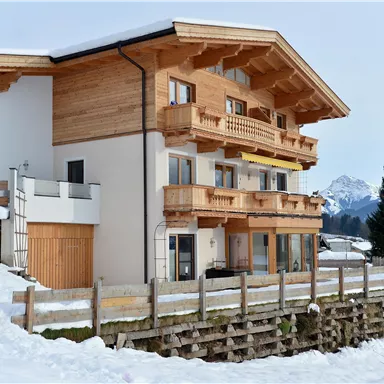 A modern chalet in the snow with wooden cladding and several balconies. Snow-covered mountains are visible in the background.