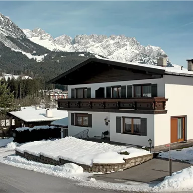 Ein modernes Haus im Schnee, umgeben von hohen Bergen. Der Himmel ist klar und die Landschaft wirkt ruhig und einladend.