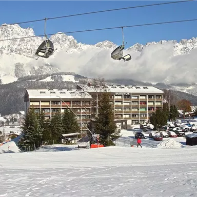 Ein schneebedecktes Bergpanorama mit einem Hotel im Vordergrund. Im Hintergrund sind die majestätischen Berge und eine Seilbahn zu sehen.