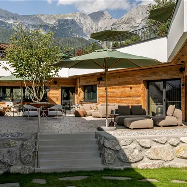 Eine moderne Terrasse mit gemütlichen Loungemöbeln und Sonnenschirmen. Im Hintergrund sind majestätische Berge zu sehen.