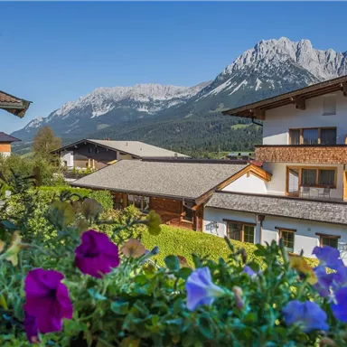 A picturesque view of a hotel with colorful flowers in the foreground and majestic mountains in the background. The sky is clear and blue.
