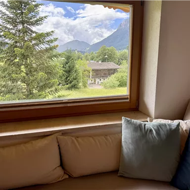 A cozy view through the window of a green landscape with trees and mountains in the background. In the foreground, soft cushions are visible on a couch.