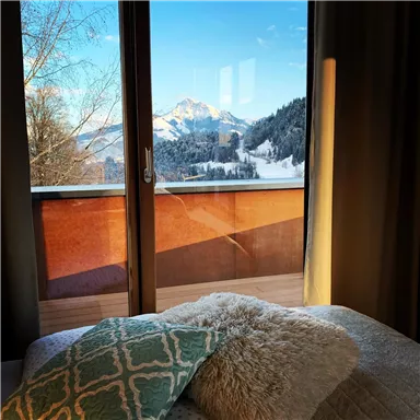 Ein Aussicht von einem gemütlichen Zimmer mit einem Bett und Kissen. Im Hintergrund sind schneebedeckte Berge und ein klarer blauer Himmel zu sehen.