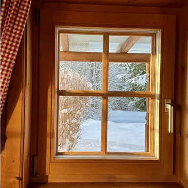 Ein Fenster mit holzrahmen, das auf eine verschneite Landschaft blickt. Die sanften Sonnenstrahlen tauchen den Raum in warmes Licht.