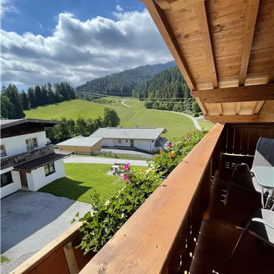 A beautiful view from a balcony overlooking a green landscape with hills and clouds. In the foreground, wooden furniture and blooming plants are visible.
