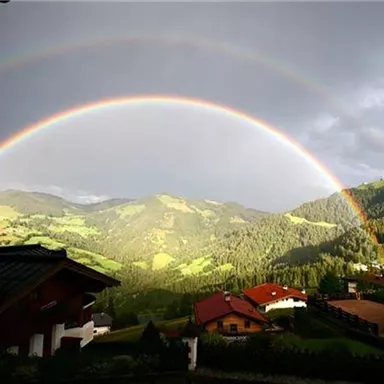 Ein schöner Regenbogen über grünen Hügeln und Hütten. Der Himmel ist teils bewölkt mit Lichtstrahlen, die die Landschaft erhellen.