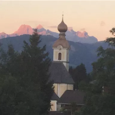 Eine malerische Kirche mit einer Zwiebelhaube vor einer beeindruckenden Berglandschaft. Die Abenddämmerung beleuchtet die Berge im Hintergrund.