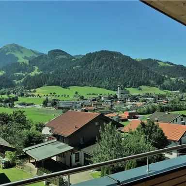 Eine malerische Landschaft mit grünen Wiesen und Bergen im Hintergrund. Im Vordergrund sind einige Häuser und eine kleine Kirche zu sehen.