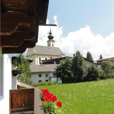 Eine malerische Landschaft mit grünen Wiesen und einer kleinen Kirche. Im Vordergrund blühen rote Geranien auf einem Balkon.