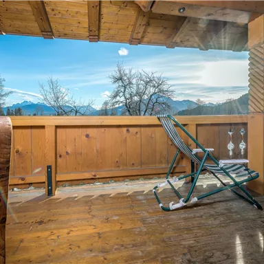 A cozy terrace with a wooden railing and a view of the mountains. The sky is clear and the sun is shining.