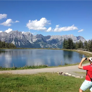 A person is photographing a picturesque mountain landscape with a clear lake in the foreground. The surroundings are green and surrounded by impressive mountains.
