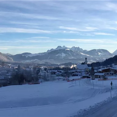 A winter landscape with snow-covered mountains and a small town in the foreground. The sky is clear and blue, emphasizing the calm atmosphere.