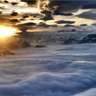 Ein Sonnenaufgang über schneebedeckten Bergen. Der Himmel ist mit dramatischen Wolken und einem Meer aus Nebel gefüllt.