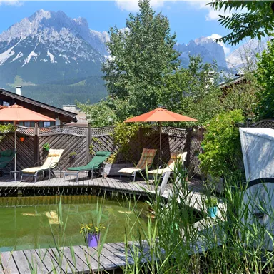 A tranquil garden with a pond and sun loungers. In the background, majestic mountains rise under a clear sky.