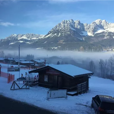 A snowy mountain landscape with a wooden house in the foreground. In the background, majestic mountains and fog are visible.