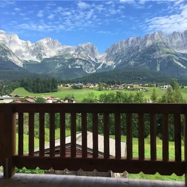 Eine beeindruckende Berglandschaft mit schneebedeckten Gipfeln und einem klaren blauen Himmel. Im Vordergrund ist eine  Balkonansicht auf grüne Wiesen und ein kleines Dorf.