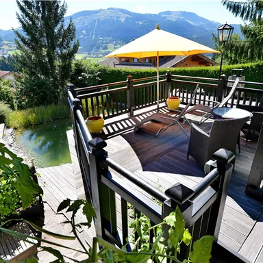 A cozy terrace with a wooden railing and garden furniture. In the background, there are mountains and a sunny sky.