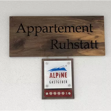 A wooden sign with the inscription "Apartment Ruhstatt" and a logo of "Alpine Hosts." The sign is mounted on a wall and depicts an inviting accommodation area.